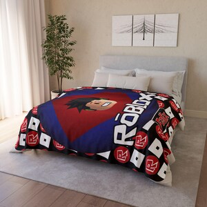 Peut inclure: Une couverture en polaire noire et rouge avec un logo Roblox et le visage d'un personnage Roblox au centre. La couverture a un motif r&eacute;p&eacute;titif du logo Roblox sur un fond noir.