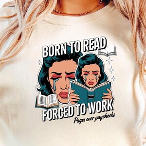 May include: Cream-colored t-shirt featuring a pop art style illustration of a crying woman reading a book. Text reads "BORN TO READ FORCED TO WORK Pages over paychecks."