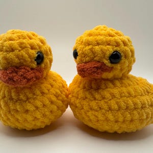 May include: Two yellow crocheted duck plushies with black eyes and orange beaks. The ducks are sitting on a white surface.