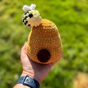 Pode incluir: Uma colmeia de crochê com uma abelha de crochê no topo. A colmeia é marrom e a abelha é amarela e preta com asas brancas.