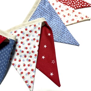 Peut inclure: Gros plan sur une guirlande de fanions color&eacute;s avec divers motifs de tissu. La guirlande pr&eacute;sente des triangles rouges, blancs et bleus avec des motifs floraux, &eacute;toil&eacute;s et g&eacute;om&eacute;triques. Les bords sont bord&eacute;s d'un ruban beige.