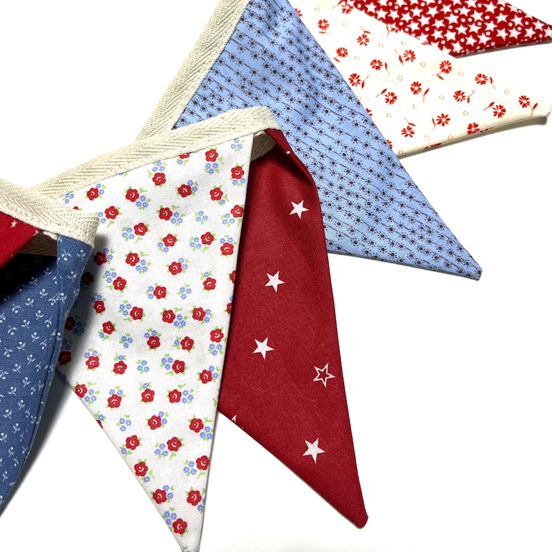 Peut inclure: Gros plan sur une guirlande de fanions color&eacute;s avec divers motifs de tissu. La guirlande pr&eacute;sente des triangles rouges, blancs et bleus avec des motifs floraux, &eacute;toil&eacute;s et g&eacute;om&eacute;triques. Les bords sont bord&eacute;s d'un ruban beige.