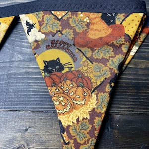 May include: Triangular Halloween bunting with a dark blue border. The fabric features a repeating pattern of pumpkins, leaves, a witch's hat, and a black cat. The words "Happy Halloween" are printed on the fabric.