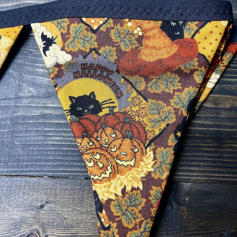 May include: Triangular Halloween bunting with a dark blue border. The fabric features a repeating pattern of pumpkins, leaves, a witch's hat, and a black cat. The words "Happy Halloween" are printed on the fabric.