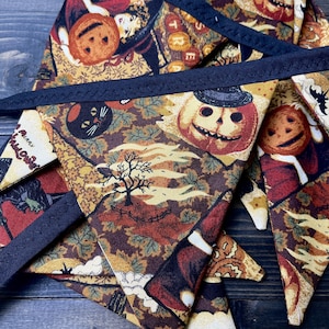 May include: A Halloween-themed banner with triangular pennants. The fabric features a repeating pattern of pumpkins, black cats, and autumn leaves in shades of orange, brown, and cream. The banner is trimmed with a dark blue border.