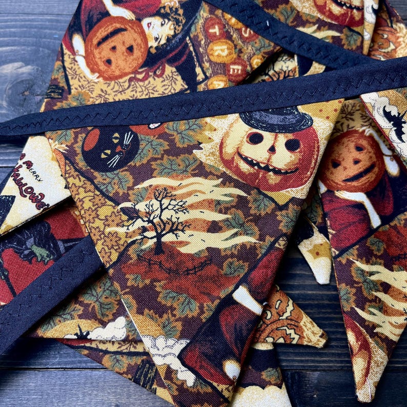 May include: A Halloween-themed banner with triangular pennants. The fabric features a repeating pattern of pumpkins, black cats, and autumn leaves in shades of orange, brown, and cream. The banner is trimmed with a dark blue border.
