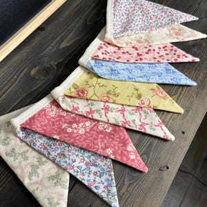 May include: A colorful fabric banner with triangular pennants. The pennants feature floral patterns in pink, blue, yellow, and cream colors. The banner is made of fabric and has a white trim.