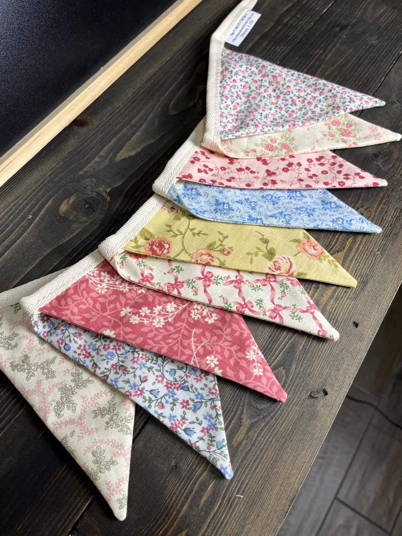 May include: A colorful fabric banner with triangular pennants. The pennants feature floral patterns in pink, blue, yellow, and cream colors. The banner is made of fabric and has a white trim.