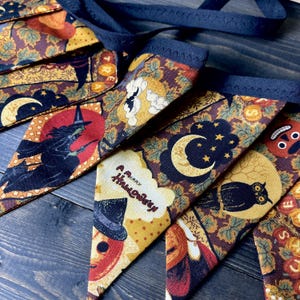 May include: A Halloween-themed banner with various festive designs. The pennants feature images of a witch, owl, moon, and pumpkin, along with the text "A Merry Halloween." The banner is made of fabric with a dark blue trim and a matching ribbon.