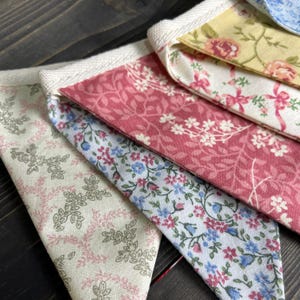 May include: Close-up of colorful fabric bunting with floral patterns. The bunting features various designs, including pink, blue, and yellow floral prints. The edges are trimmed with white fabric.