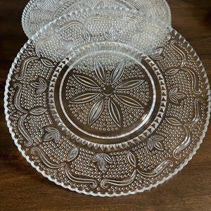 May include: A stack of clear glass plates with intricate embossed floral and dot patterns. The plates have a scalloped edge and a central starburst design. The plates are arranged on a dark wooden surface.