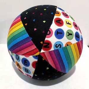 Op de afbeelding: Een veelkleurige stoffen bal met een regenboogstrepenpatroon, een zwart stoffen paneel met kleine roze hartjes en een wit stoffen paneel met kleurrijke cirkels met letters van het alfabet.
