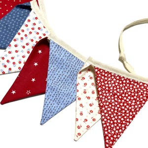 Peut inclure: Guirlande de fanions en tissu rouge, blanc et bleu. Les fanions triangulaires pr&eacute;sentent des motifs vari&eacute;s : &eacute;toiles, motifs floraux et petits motifs g&eacute;om&eacute;triques. La guirlande est bord&eacute;e d'un ruban cr&egrave;me.