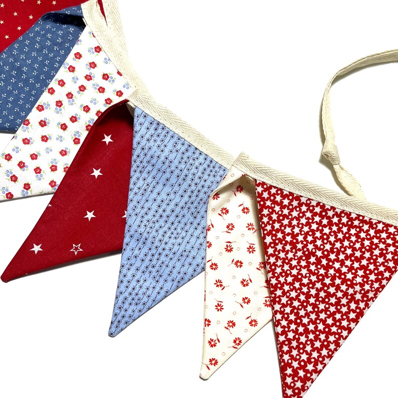 Peut inclure: Guirlande de fanions en tissu rouge, blanc et bleu. Les fanions triangulaires pr&eacute;sentent des motifs vari&eacute;s : &eacute;toiles, motifs floraux et petits motifs g&eacute;om&eacute;triques. La guirlande est bord&eacute;e d'un ruban cr&egrave;me.