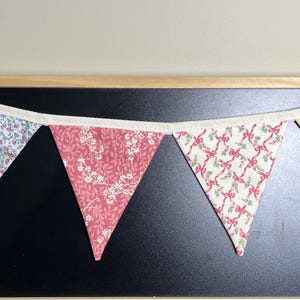 May include: A colorful fabric banner with triangular flags. The flags display various floral patterns in shades of pink, blue, yellow, and white. The banner is strung on a white ribbon and is set against a black background.