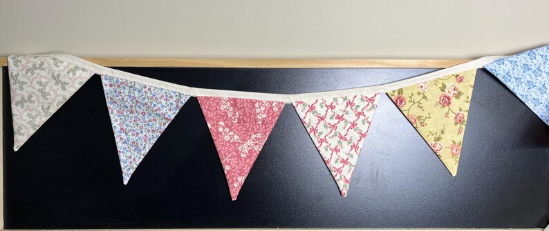 May include: A colorful fabric banner with triangular flags. The flags display various floral patterns in shades of pink, blue, yellow, and white. The banner is strung on a white ribbon and is set against a black background.