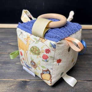 May include: A multi-textured baby toy cube with a variety of fabrics and textures. The cube features a deer, turtle, owl, and other woodland creatures. The cube has a wooden ring for teething and colorful ribbons for sensory play.