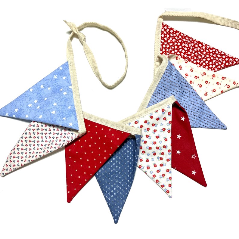 Peut inclure: Une guirlande de fanions patriotiques avec des drapeaux triangulaires rouges, blancs et bleus. Les fanions pr&eacute;sentent des motifs d'&eacute;toiles et de fleurs, avec une bordure cr&egrave;me et un ruban assorti pour l'accrochage. Parfait pour les d&eacute;corations.
