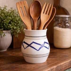 May include: A white ceramic utensil holder with blue diamond pattern holds wooden spoons, a spatula, and a fork. The holder sits on a dark wooden countertop, with a small potted plant and a jar in the background.