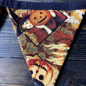 May include: Triangular Halloween banner with a dark blue border. The fabric features a vintage-style print of a witch holding a jack-o'-lantern, with autumn leaves and a smiling pumpkin. The color palette includes shades of brown, orange, and yellow.