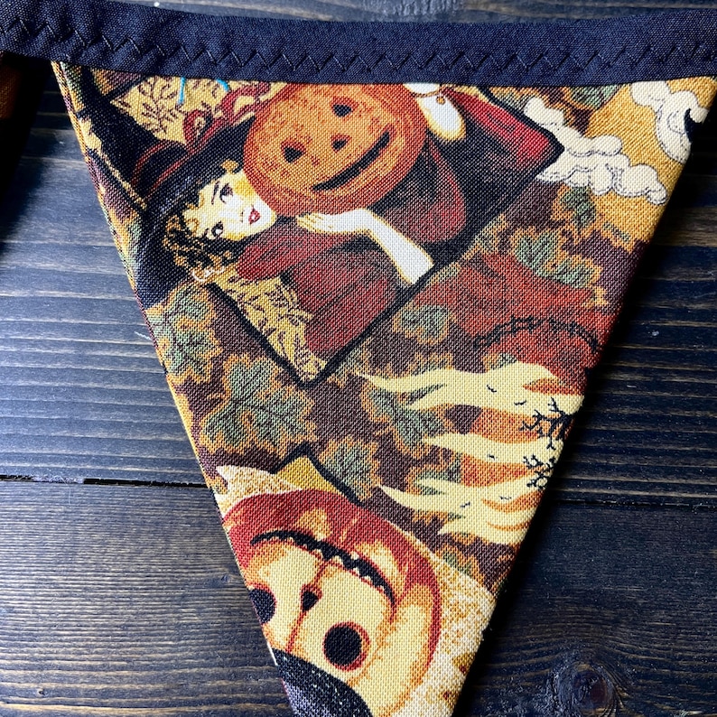 May include: Triangular Halloween banner with a dark blue border. The fabric features a vintage-style print of a witch holding a jack-o'-lantern, with autumn leaves and a smiling pumpkin. The color palette includes shades of brown, orange, and yellow.