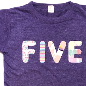 Op de afbeelding: Paars T-shirt met het woord "FIVE" in kleurrijke letters met een regenboog-, eenhoorn- en ballonpatroon.