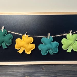 May include: A St. Patrick's Day banner featuring six felt shamrocks in shades of green and yellow, strung on twine with small wooden clothespins. The banner is displayed against a black background.
