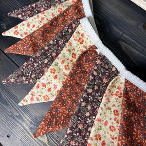 May include: A fabric banner with triangular pennants in various shades of brown and cream, adorned with floral patterns. The pennants are strung together with white ribbon, creating a rustic and charming decorative piece.