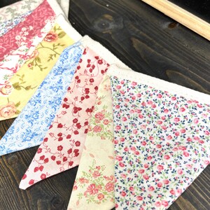 May include: Close-up of a colorful fabric bunting with a variety of floral patterns. The pennants feature designs in shades of pink, yellow, and blue, with white lace trim. The bunting is arranged on a dark wooden surface.