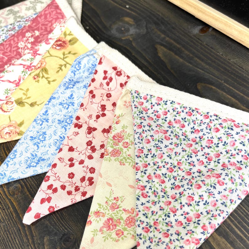 May include: Close-up of a colorful fabric bunting with a variety of floral patterns. The pennants feature designs in shades of pink, yellow, and blue, with white lace trim. The bunting is arranged on a dark wooden surface.
