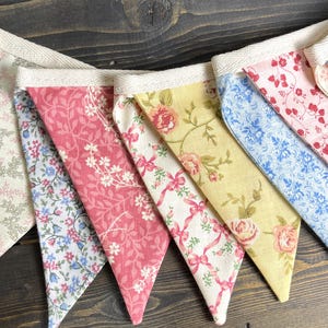 May include: A colorful fabric banner with triangular pennants. The pennants feature floral patterns in shades of pink, blue, yellow, and white, strung together with a cream-colored ribbon. The banner is perfect for adding a touch of charm to any space.