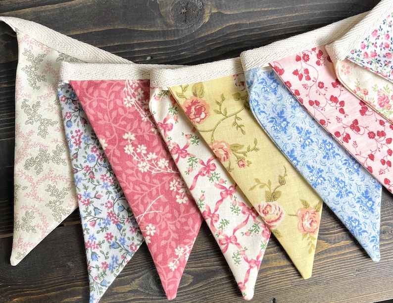 May include: A colorful fabric banner with triangular pennants. The pennants feature floral patterns in shades of pink, blue, yellow, and white, strung together with a cream-colored ribbon. The banner is perfect for adding a touch of charm to any space.