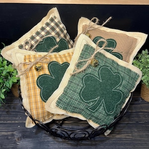 以下が含まれることがあります： 黒い金属製のバスケットに入った、クローバー型の装飾用枕のコレクション。枕は、チェック柄の生地に緑色のクローバーが特徴で、ジュートの縁と紐のアクセントが付いています。各枕には小さな金のベルが付いています。