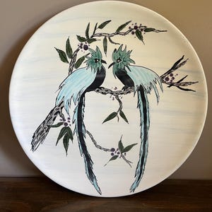 May include: A white ceramic plate with a hand-painted design of two stylized blue birds perched on a branch with green leaves and purple berries. The birds have long, flowing tail feathers. The plate has a light blue, textured background.