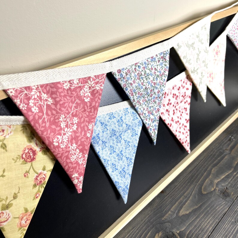 May include: A colorful bunting banner with triangular fabric flags. The flags feature floral patterns in shades of pink, blue, yellow, and white. The banner is attached to a white ribbon and is displayed against a black background.