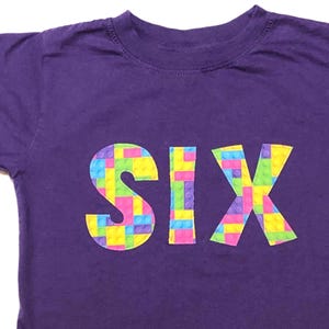 May include: Purple t-shirt with the word "SIX" spelled out in colorful plastic building blocks.