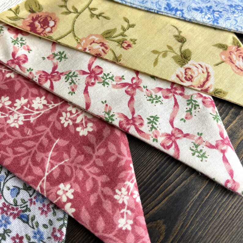 May include: Close-up of several fabric swatches with floral patterns. The fabrics feature designs in shades of pink, yellow, and blue. One swatch has pink bows and green accents. Another has pink roses on a yellow background.