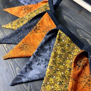 May include: A close-up of a Halloween-themed banner with triangular flags. The flags feature various patterns, including spiderwebs, floral designs, and polka dots, in orange, yellow, and navy blue. The banner is trimmed with black fabric.