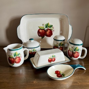 May include: A ceramic kitchen set featuring a cream-colored base with a dark green rim and a red apple motif. The collection includes a rectangular serving platter, a lidded sugar bowl, a creamer, a butter dish, two shakers, and a spoon rest.