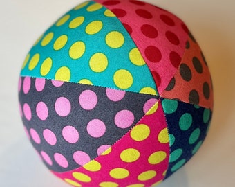May include: A multi-colored fabric ball with a polka dot pattern. The ball is made of six different fabric panels, each with a different color and polka dot pattern. The colors are teal, pink, red, gray, dark blue, and yellow.