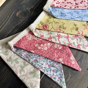 May include: A colorful fabric banner with a variety of floral patterns in pink, blue, and yellow. The banner is made of triangular fabric pieces sewn together with a white trim. The banner is displayed on a dark wooden surface.