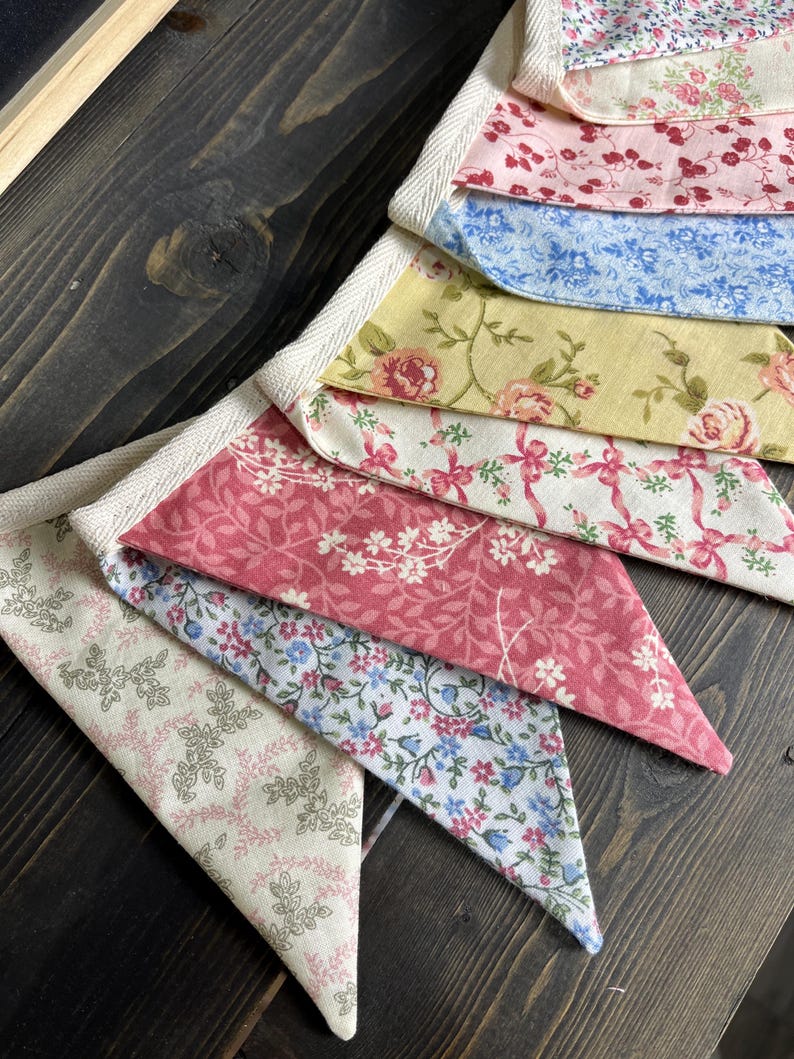 May include: A colorful fabric banner with a variety of floral patterns in pink, blue, and yellow. The banner is made of triangular fabric pieces sewn together with a white trim. The banner is displayed on a dark wooden surface.