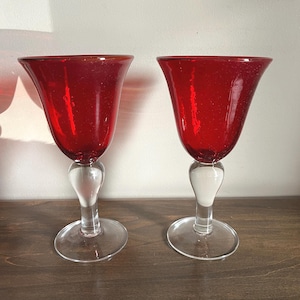 May include: Two matching goblets featuring deep red, translucent glass bowls with a subtle bubbled texture. Each goblet has a clear, smooth glass stem with a rounded bulbous accent near the base and a wide, flat, circular clear glass foot.