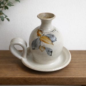 May include: A ceramic candle holder with a round body, a small handle, and a wide base. The off-white piece features a floral design in muted tones of gray, yellow, and black. The candle holder sits on a wooden surface.