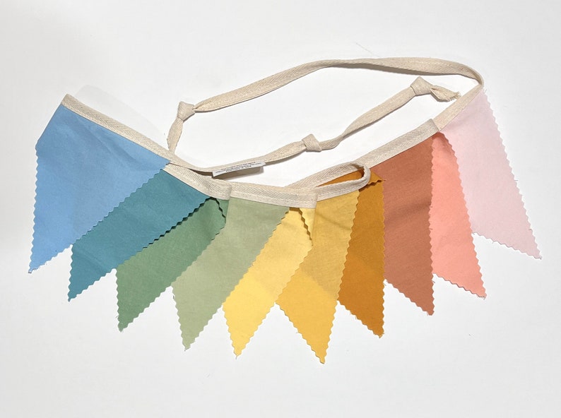 Puede incluir: Una pancarta de tela de colores arco&iacute;ris con bordes festoneados. La pancarta est&aacute; hecha de tela suave y natural y presenta una variedad de colores, incluyendo azul, verde, amarillo, naranja y rosa. La pancarta est&aacute; enhebrada con un cord&oacute;n de algod&oacute;n blanco.