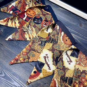 May include: A collection of Halloween-themed triangular bandanas. The bandanas feature a repeating pattern of pumpkins, black cats, and fall foliage in shades of brown, orange, and yellow. The text "A Merry Halloween" is also visible on some of the bandanas.