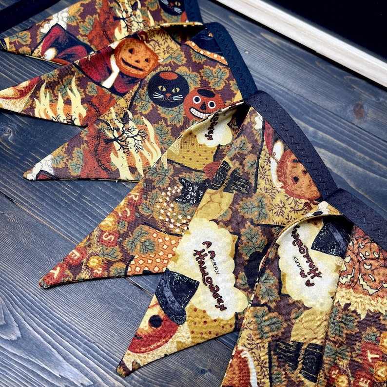 May include: A collection of Halloween-themed triangular bandanas. The bandanas feature a repeating pattern of pumpkins, black cats, and fall foliage in shades of brown, orange, and yellow. The text "A Merry Halloween" is also visible on some of the bandanas.