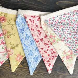 May include: A colorful fabric bunting with various floral patterns. The pennants feature designs in yellow, pink, and blue, with roses and other floral motifs. The bunting is attached to a cream-colored fabric strip.