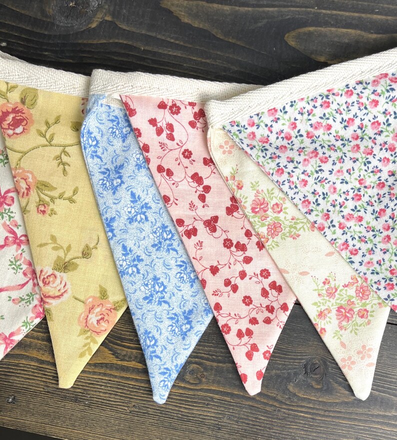May include: A colorful fabric bunting with various floral patterns. The pennants feature designs in yellow, pink, and blue, with roses and other floral motifs. The bunting is attached to a cream-colored fabric strip.
