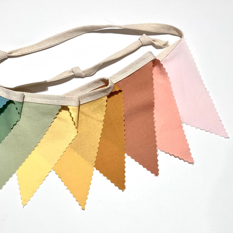 Puede incluir: Una pancarta de tela de colores arco&iacute;ris con una cuerda de algod&oacute;n blanca. La pancarta est&aacute; hecha de ocho banderas triangulares en tonos de azul, verde, amarillo, naranja, marr&oacute;n, rosa y rosa claro.